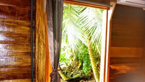 A wooden cabin room with a bed, a small table with a remote control, a decorative shell, and a window with a curtain overlooking a tropical garden. - El Jaguar (Bastimentos)