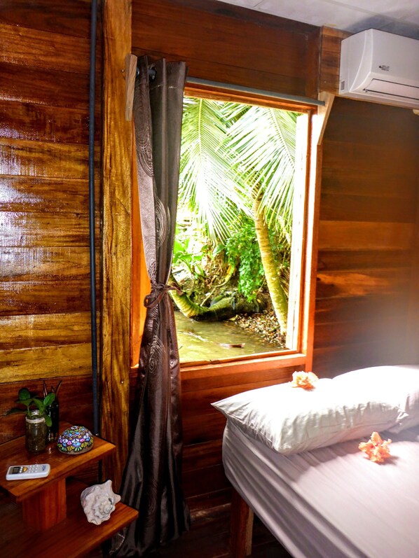Une chambre dans un chalet en bois, avec un lit, une petite table sur laquelle se trouve une télécommande, un coquillage décoratif et une fenêtre avec un rideau donnant sur un jardin tropical.