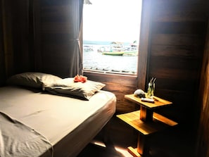 Una habitación con cama, ventana con vista a un cuerpo de agua y una pequeña mesa de madera con un control remoto y un objeto decorativo.