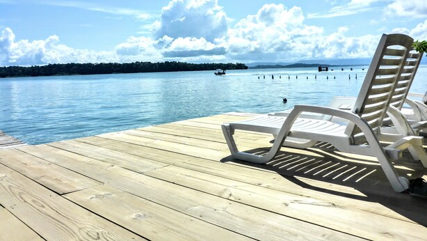 A wooden deck with white lounge chairs facing a calm lake, surrounded by trees and a clear sky. - El Jaguar (Bastimentos)