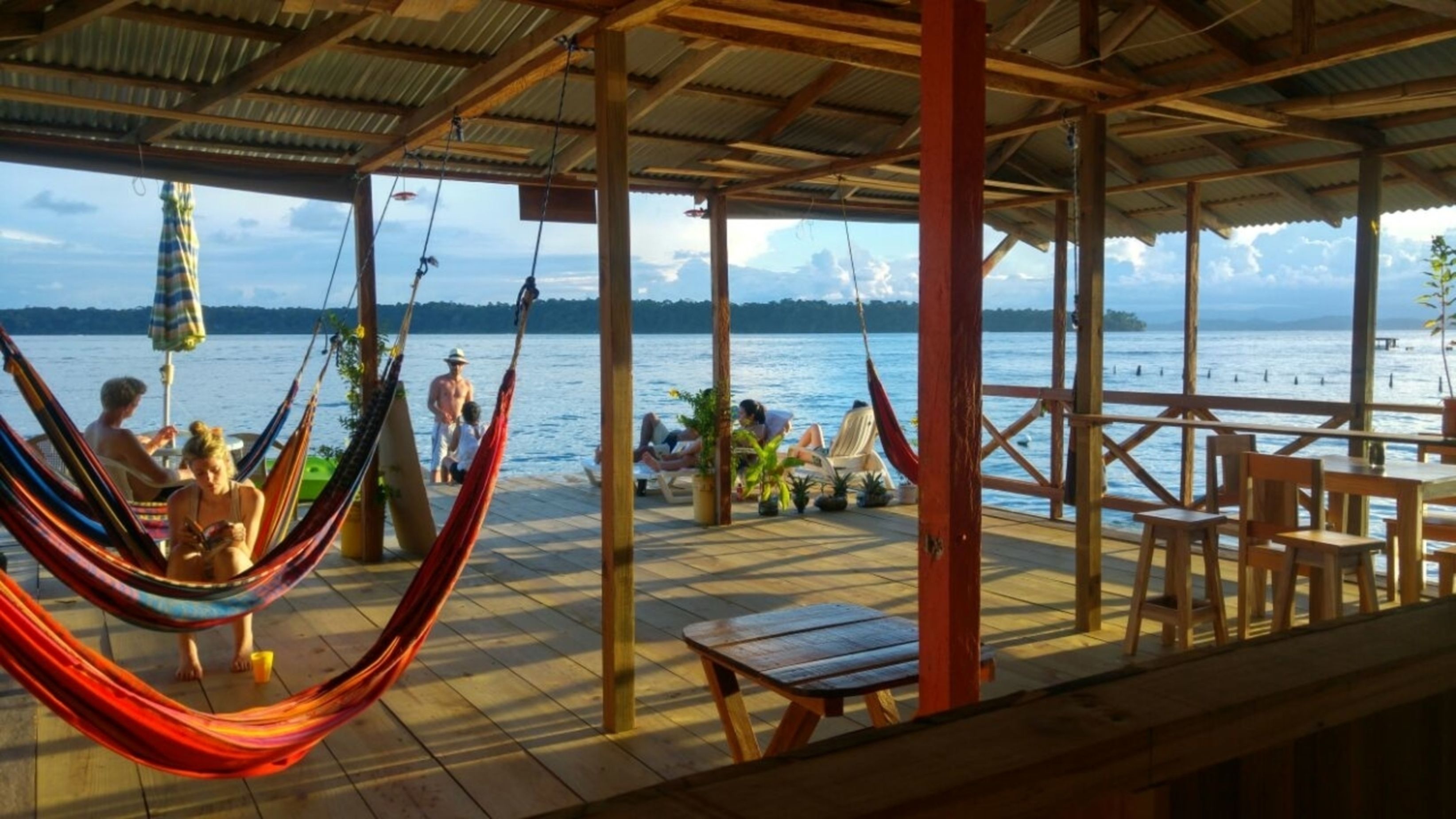 A covered outdoor area with wooden tables and chairs, a hammock, and a view of the water.