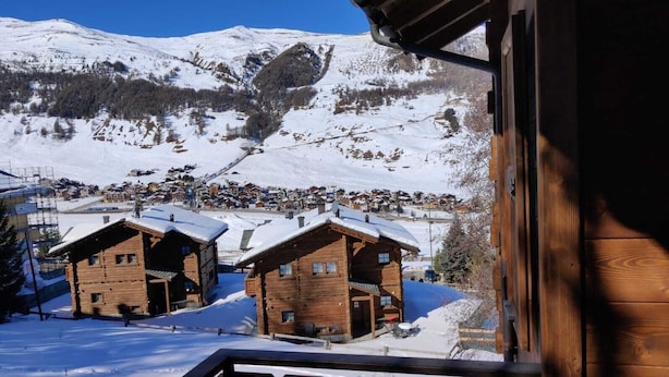 Una cabaña de madera con techo nevado, con una montaña nevada y un pequeño pueblo al fondo.