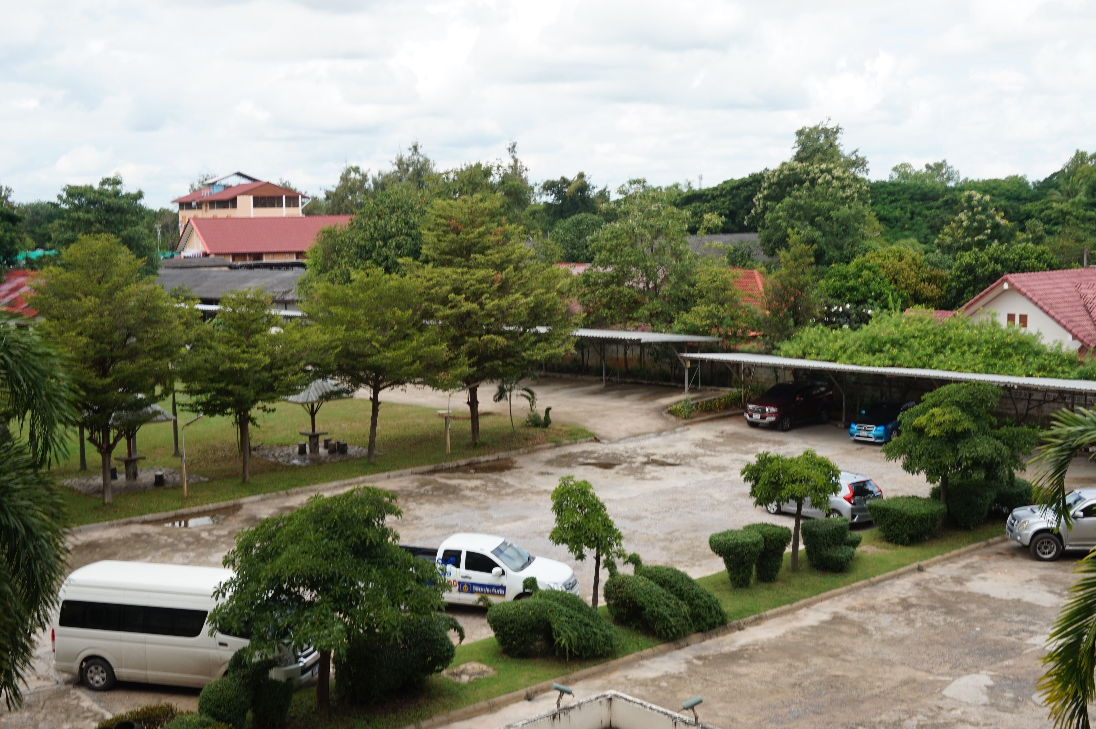 A parking area with several cars and a white van, surrounded by trees and a building with a red roof.