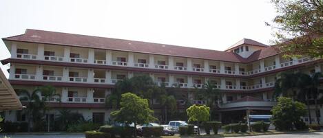 A multi-story building with a red-tiled roof, multiple balconies, and a parking area with cars and greenery.