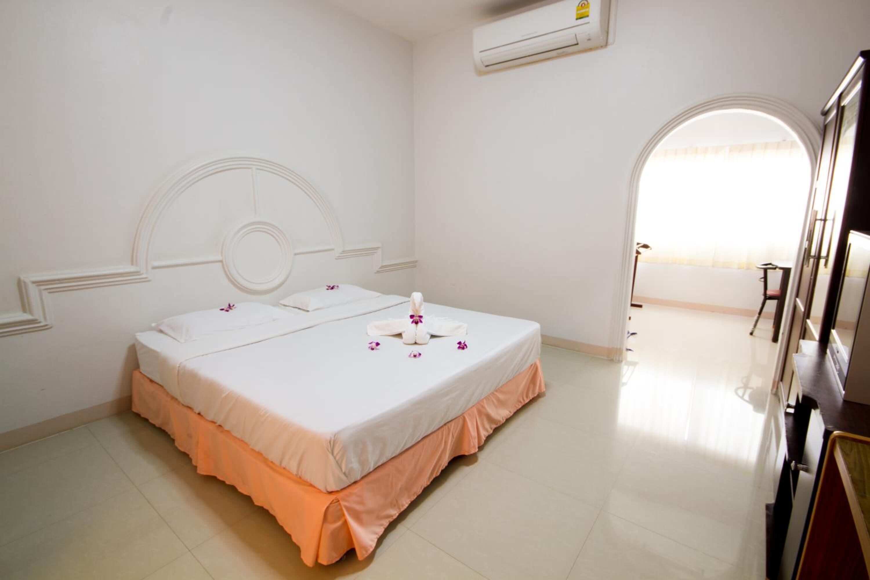 A hotel room with a double bed, a small table, an air conditioning unit, and a view of the outside through an arched window.