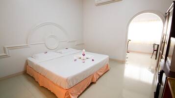 A hotel room with a double bed, a small table, an air conditioning unit, and a view of the outside through an arched window.