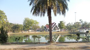 A park with a pond, palm trees, and buildings in the background. - A.P. Garden Hotel (Kalasin)