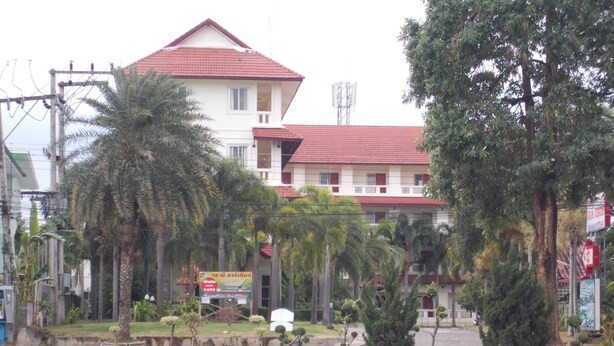 A building with a red roof and white walls, surrounded by palm trees and a body of water. - A.P. Garden Hotel (Kalasin)