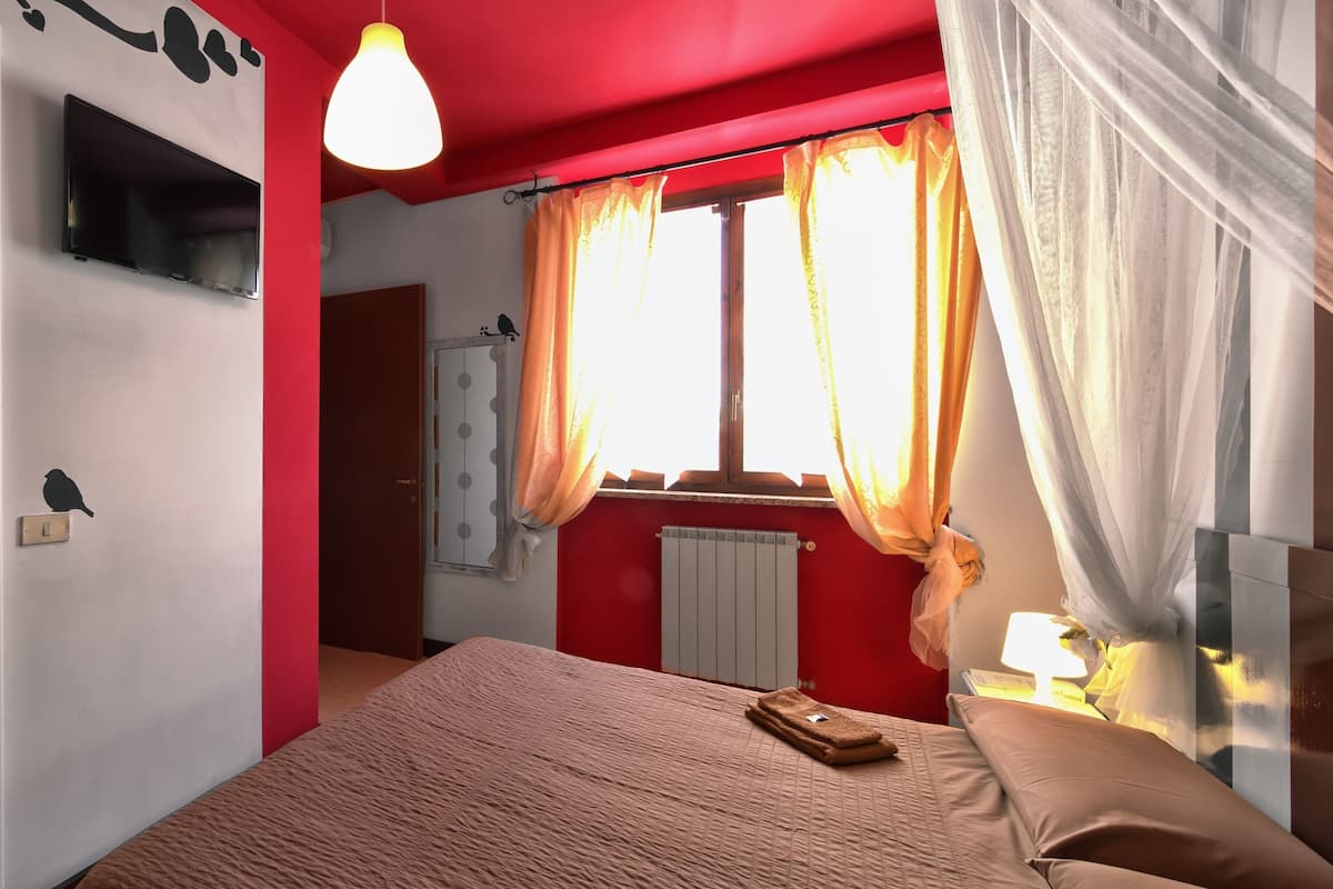 Un dormitorio con cama, mesita de noche, lámpara, ventana con cortinas, radiador y televisor montado en la pared.