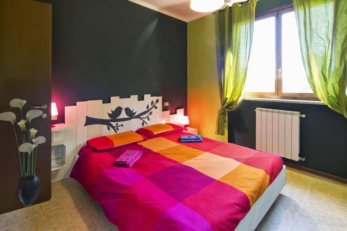 Un dormitorio con una cama, una mesita de noche, una lámpara, un radiador y una ventana con cortinas.