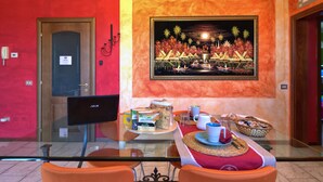 A hotel lobby with a red wall, a framed picture of a tropical scene, a reception desk with a computer, and a breakfast setup with coffee and snacks.