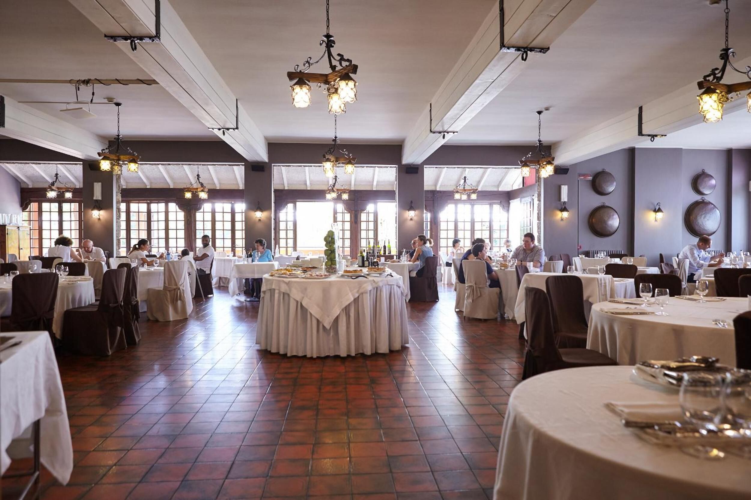 A spacious dining area with multiple round tables, white tablecloths, and hanging pendant lights.