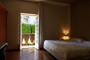 Un dormitorio con cama, mesita de noche, lámpara y una puerta abierta que da a un balcón con plantas.