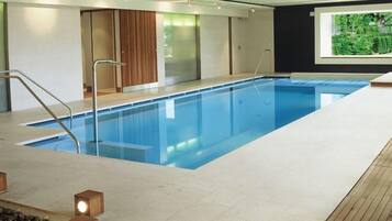 Una piscina interior moderna con paredes de vidrio transparente, una zona de cubierta de madera y vistas a la vegetación a través de una ventana.