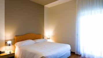 Quarto de hotel com uma cama grande, duas luminárias de cabeceira, um criado-mudo e uma janela com cortinas leves.