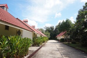 A row of single-story houses with red-tiled roofs, a concrete pathway, and lush greenery.