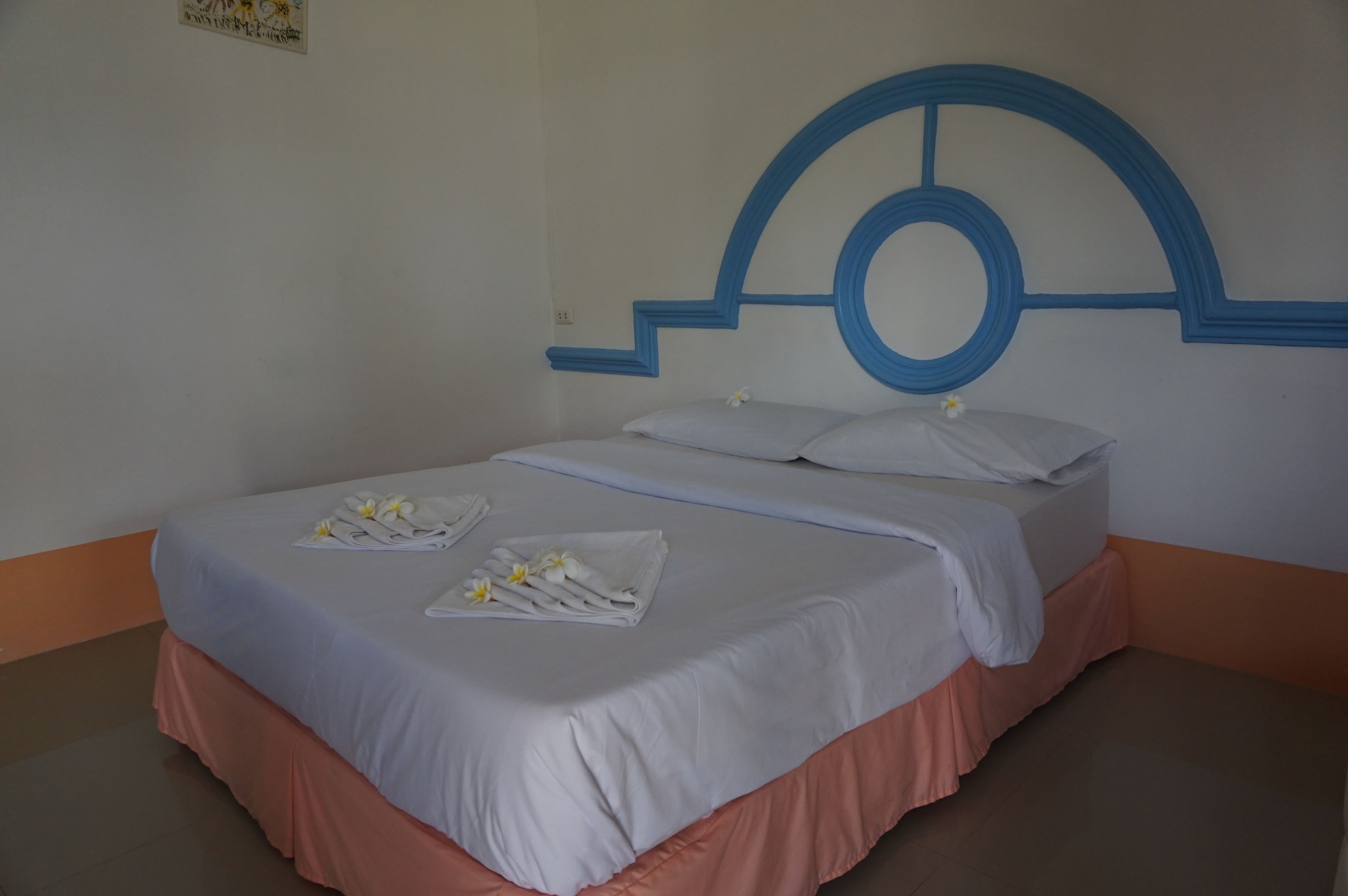 A neatly made bed with white linens and a peach-colored skirt, a blue headboard with a circular design, and a wall-mounted sign.