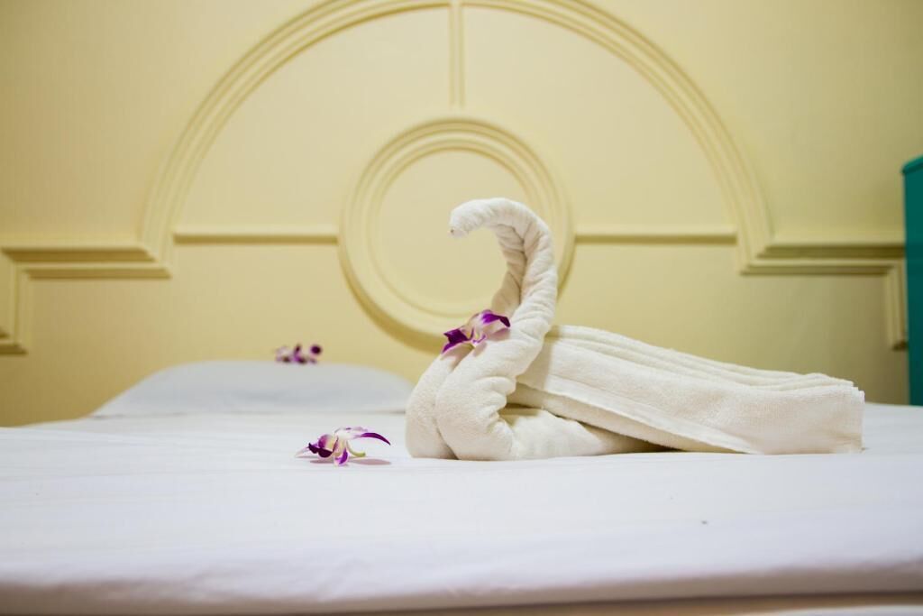 A neatly made bed with a towel shaped like a swan and a single purple orchid petal on the pillow.