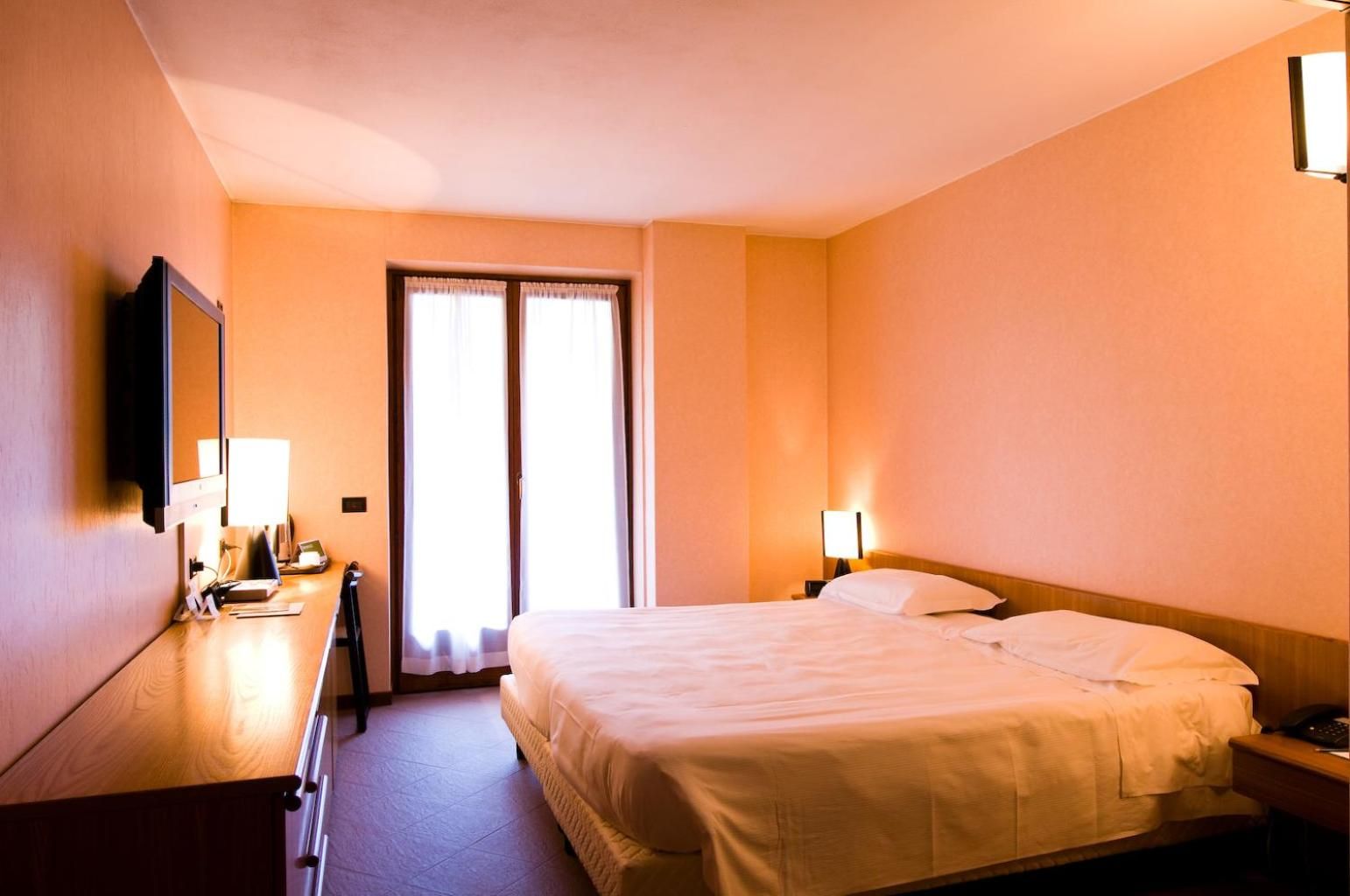 Habitación de hotel con cama, mesitas de noche, escritorio, silla, televisor y ventana con cortinas.