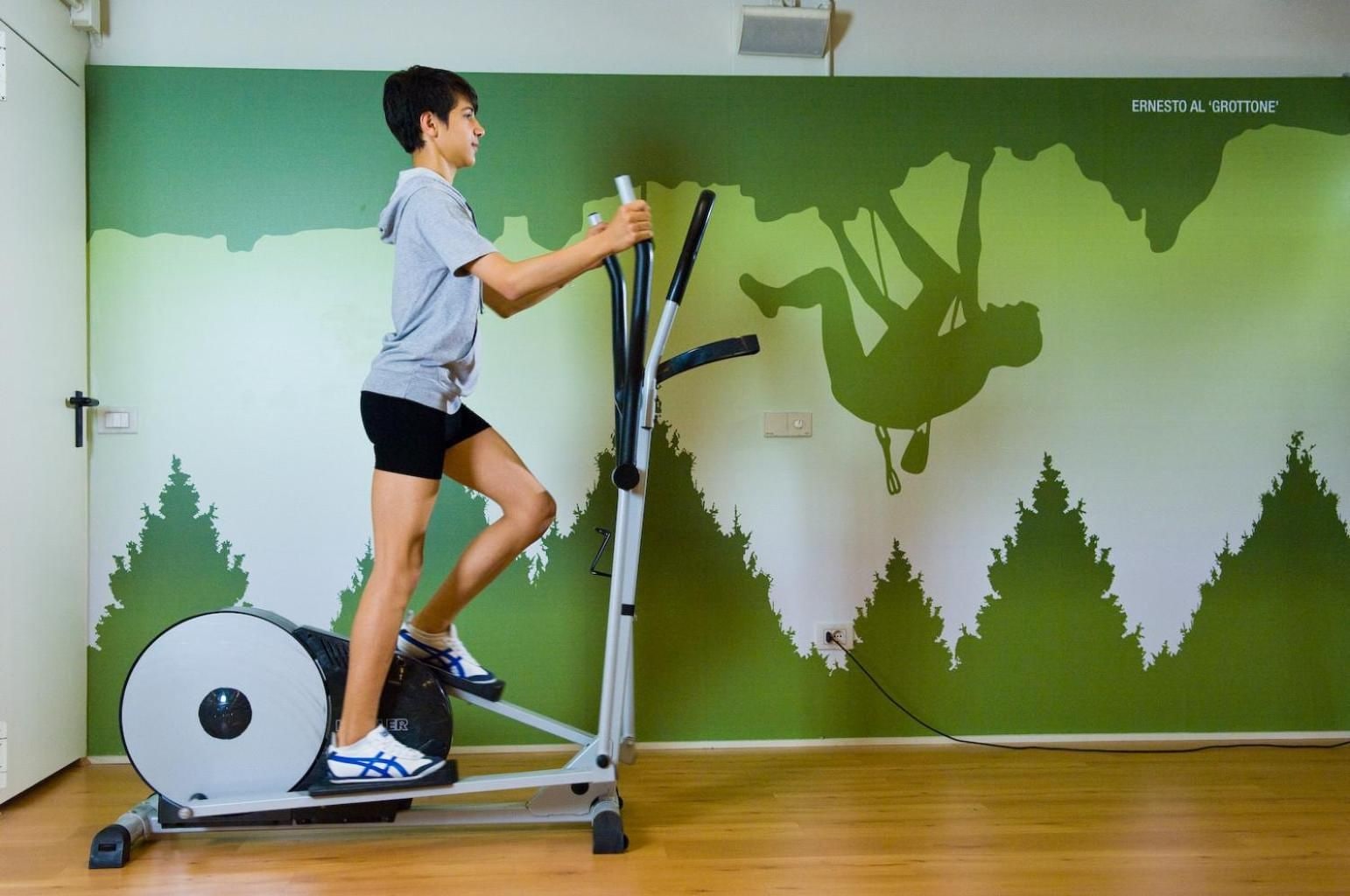 A person using an elliptical machine in a room with a green wall and a mural of trees.