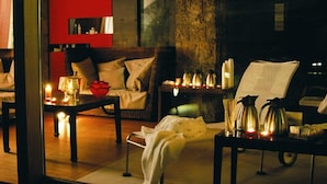 Une pièce chaleureuse avec un canapé, une table basse et un fauteuil. Il y a des bougies sur la table et une fleur rouge sur le canapé.
