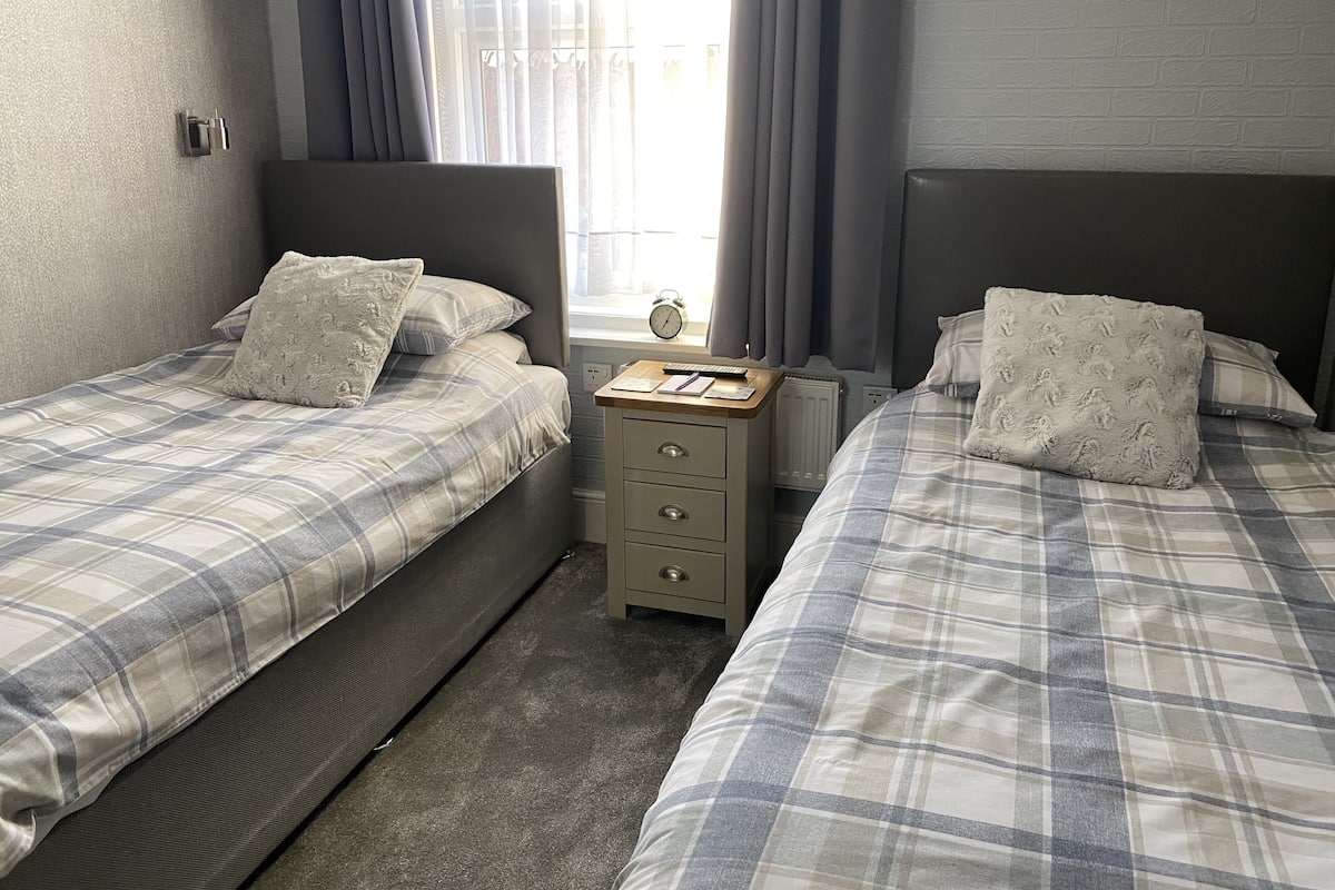 A hotel room with two beds, a nightstand, a small table, and a window with curtains.