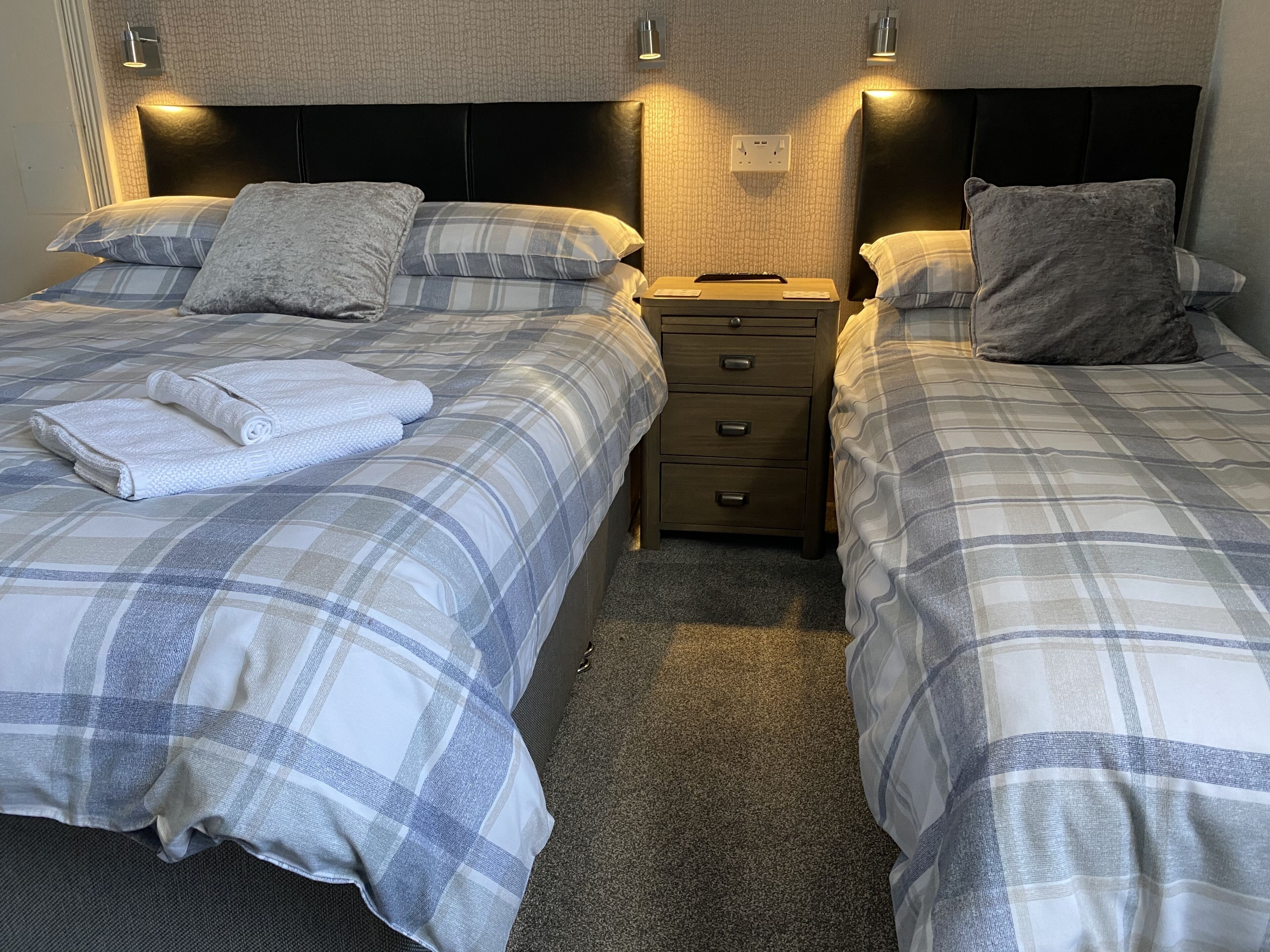 A hotel room with two beds, each with a grey and white checkered duvet, a small bedside table with a lamp, and a headboard with a textured design.