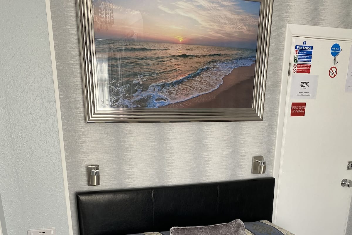 A neatly made bed with a patterned bedspread, a remote control, and a smartphone on a wooden nightstand.