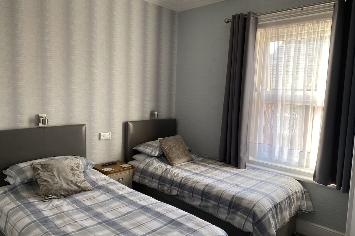 A bedroom with two beds, a nightstand, a window with curtains, and a wall-mounted light switch.