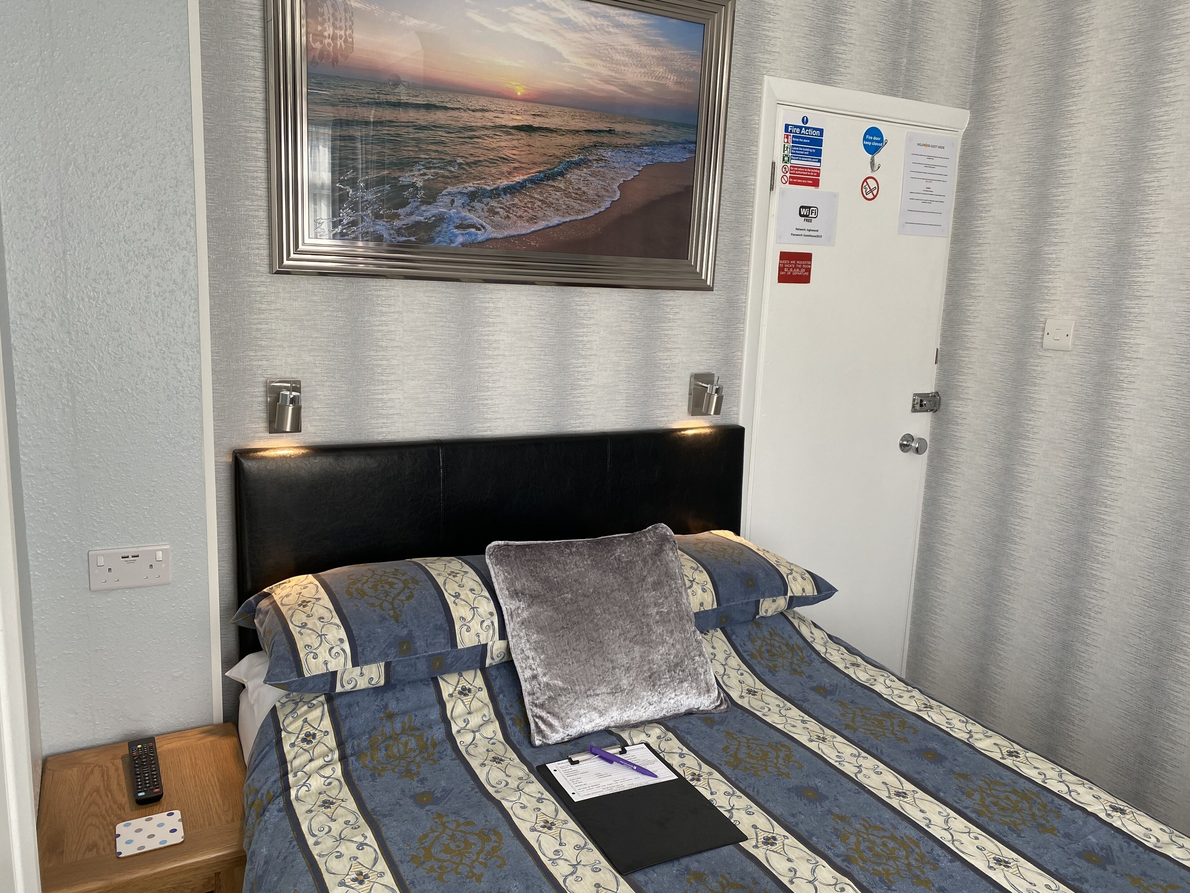 A neatly arranged hotel room with a bed, a nightstand, a remote control, a notebook, and a pen.