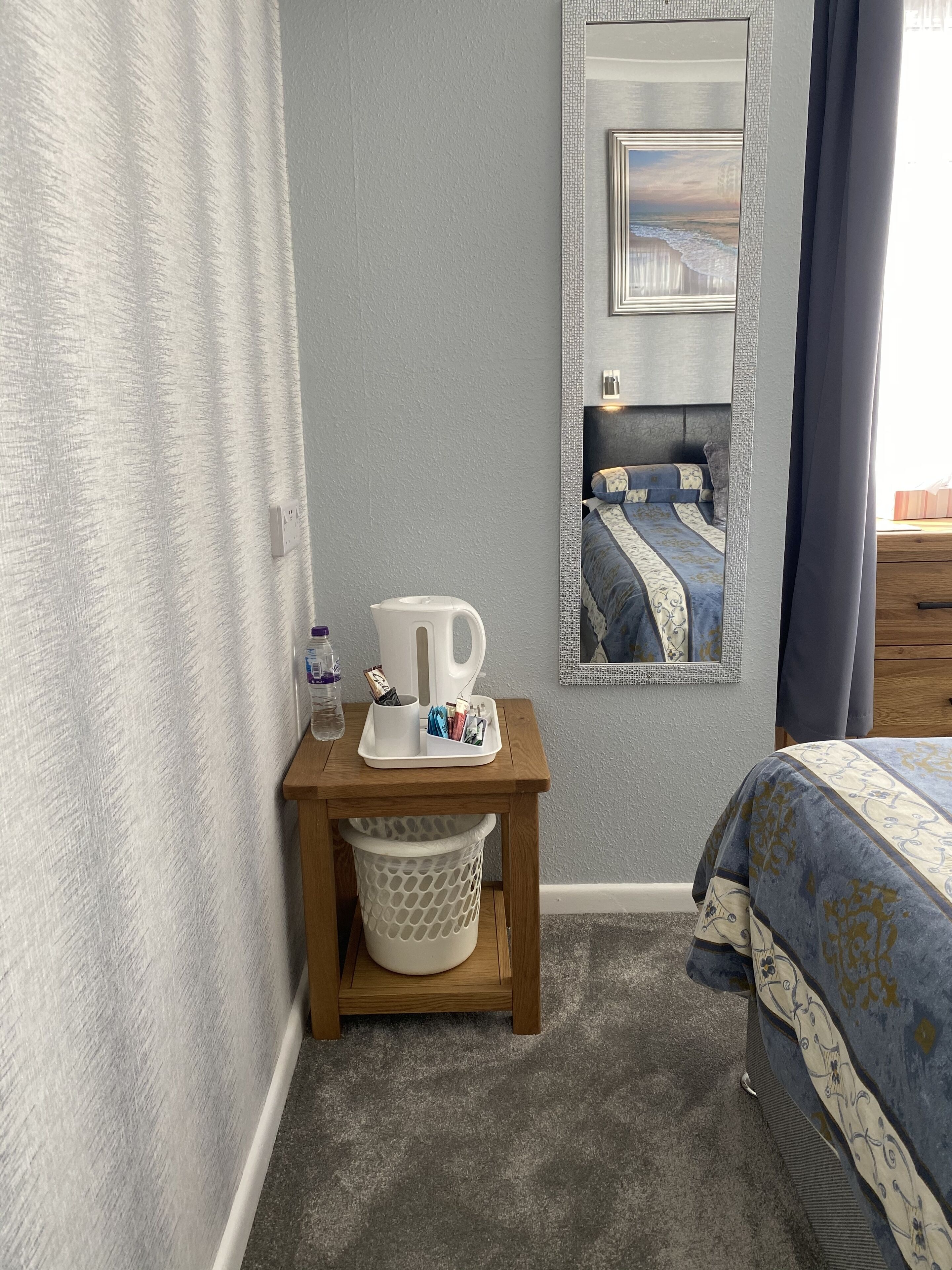 A bedroom with a bed, a bedside table with a kettle, a trash bin, and a mirror reflecting a bed with a blue and white patterned bedspread.