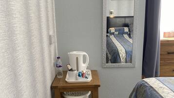 A bedroom with a bed, a bedside table with a kettle, a trash bin, and a mirror reflecting a bed with a blue and white patterned bedspread.