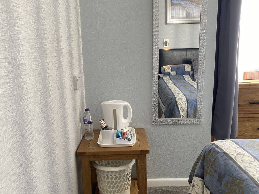 A bedroom with a bed, a bedside table with a kettle, a trash bin, and a mirror reflecting a bed with a blue and white patterned bedspread.