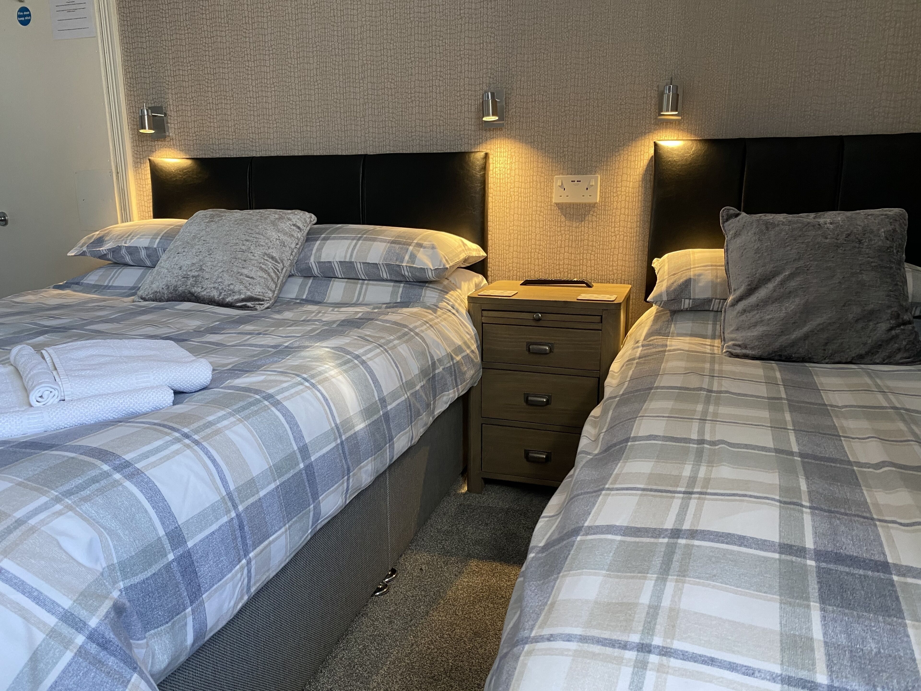 A double bed with a plaid bedspread, a bedside table with a lamp, and a wall-mounted light fixture.