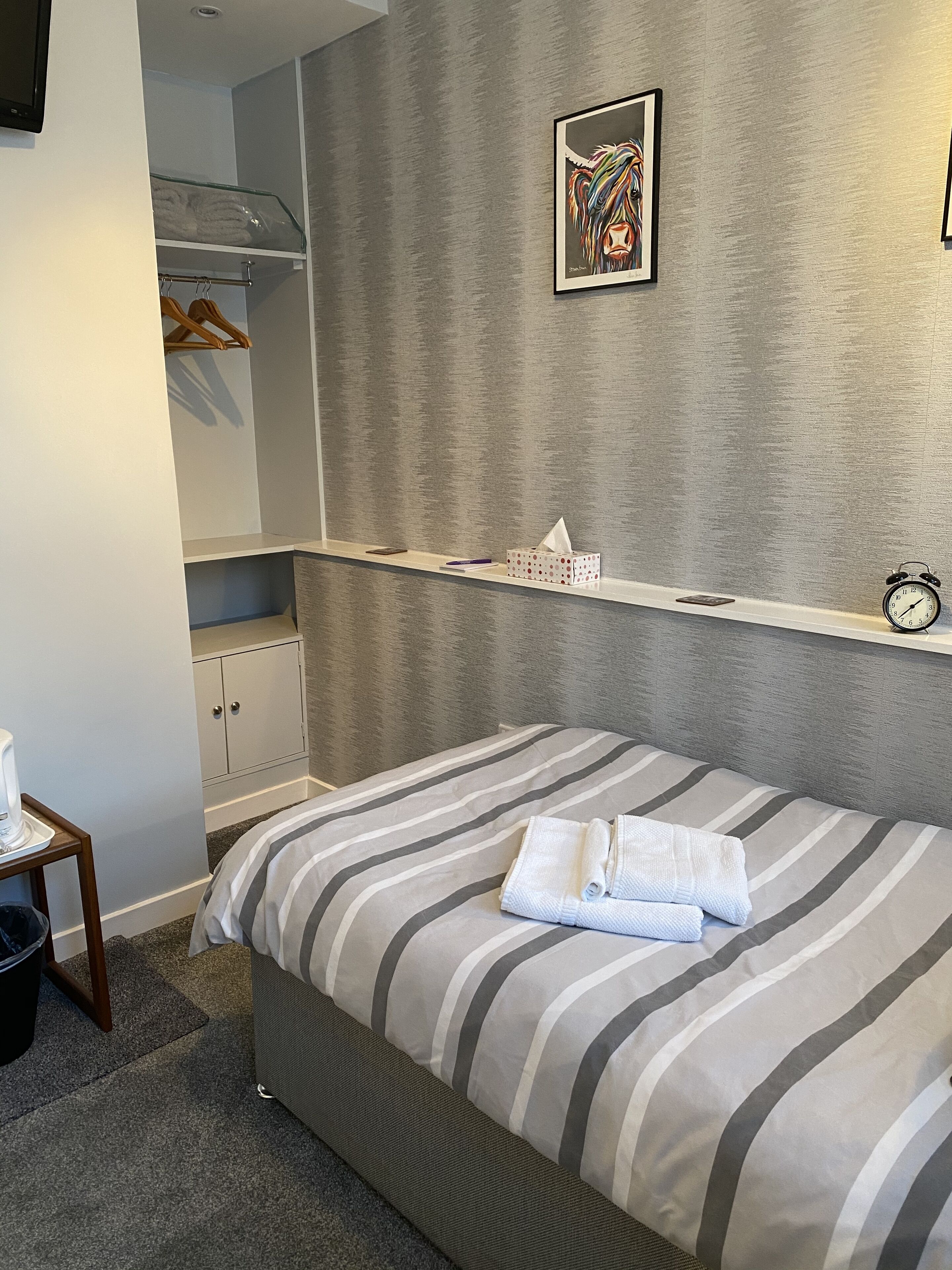 Un dormitorio ordenado con cama, mesita de noche, estante con un reloj, armario y un cuadro en la pared.