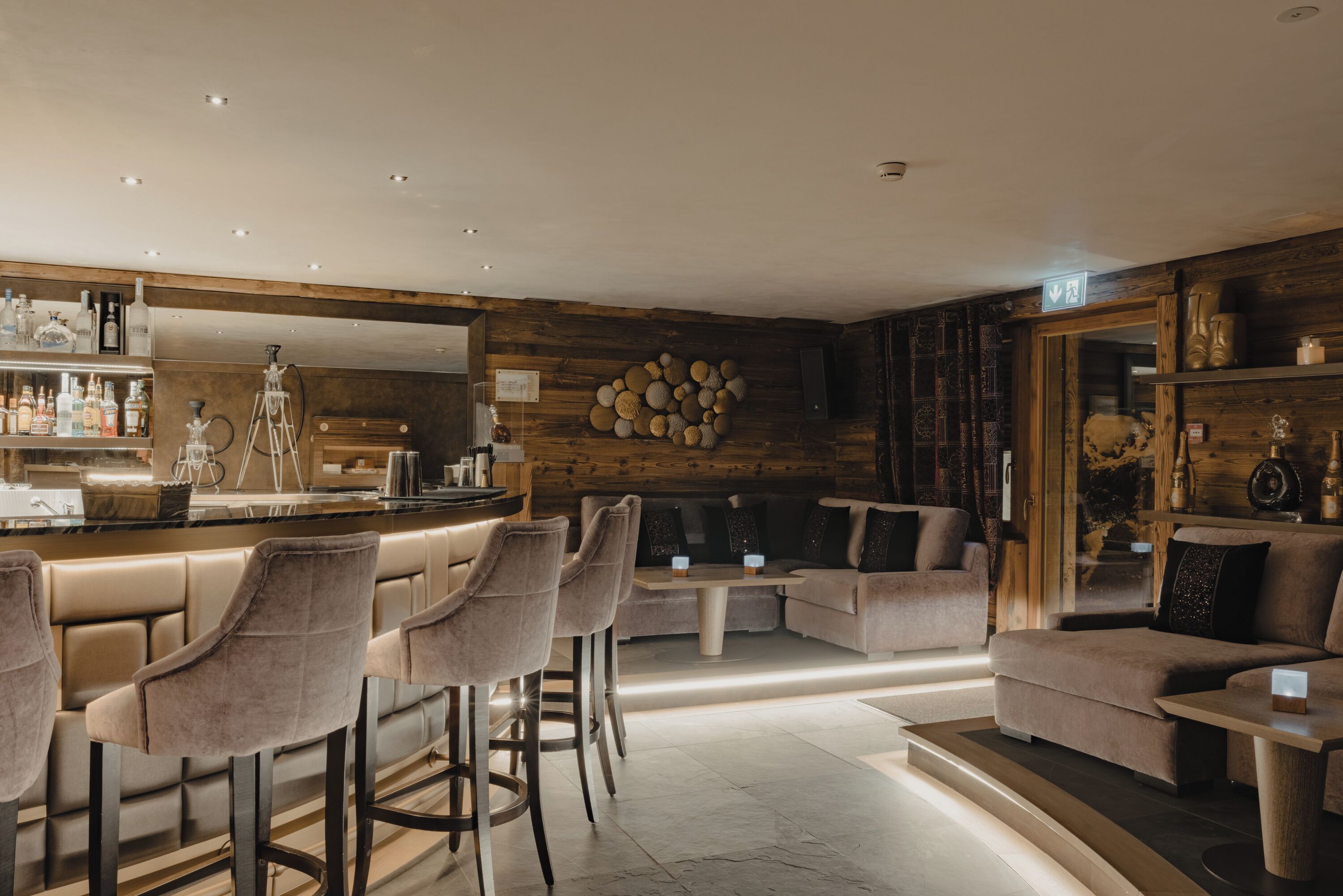 A modern bar area with bar stools, a counter with bottles, and a seating area with sofas and a coffee table.