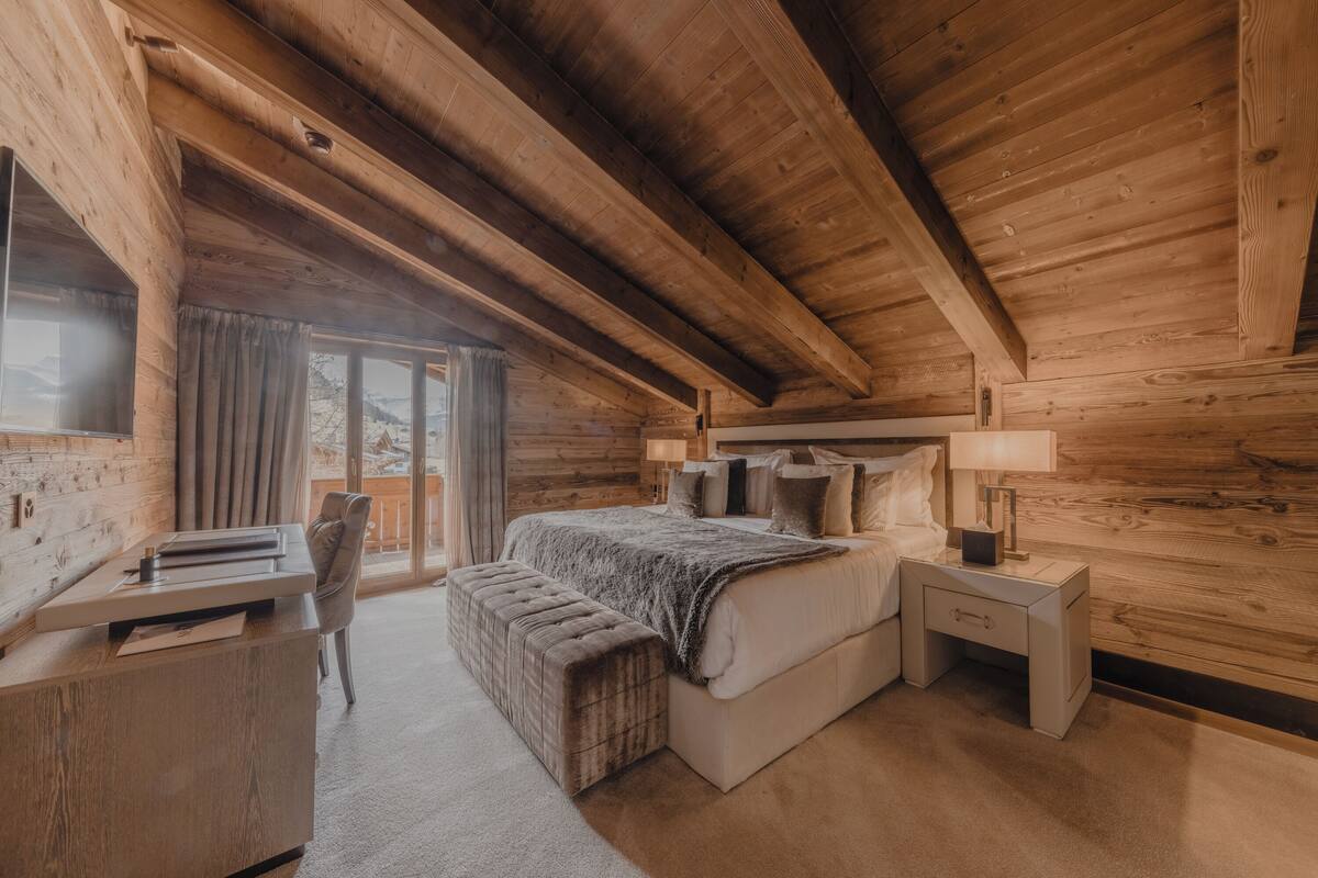 A wooden cabin bedroom with a bed, bedside tables, a desk, and a window with curtains.