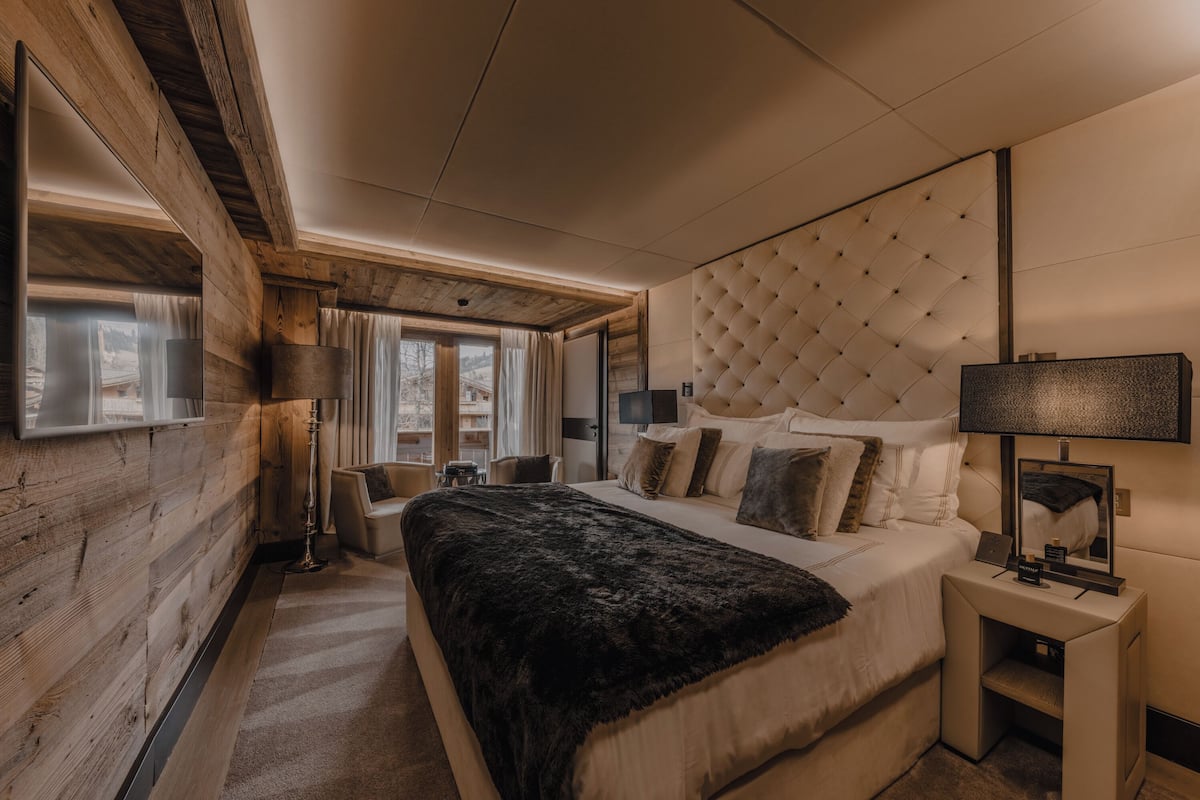 A modern bedroom with a large bed, a tufted headboard, a bedside table with a lamp, and a view of the outdoors through large windows.