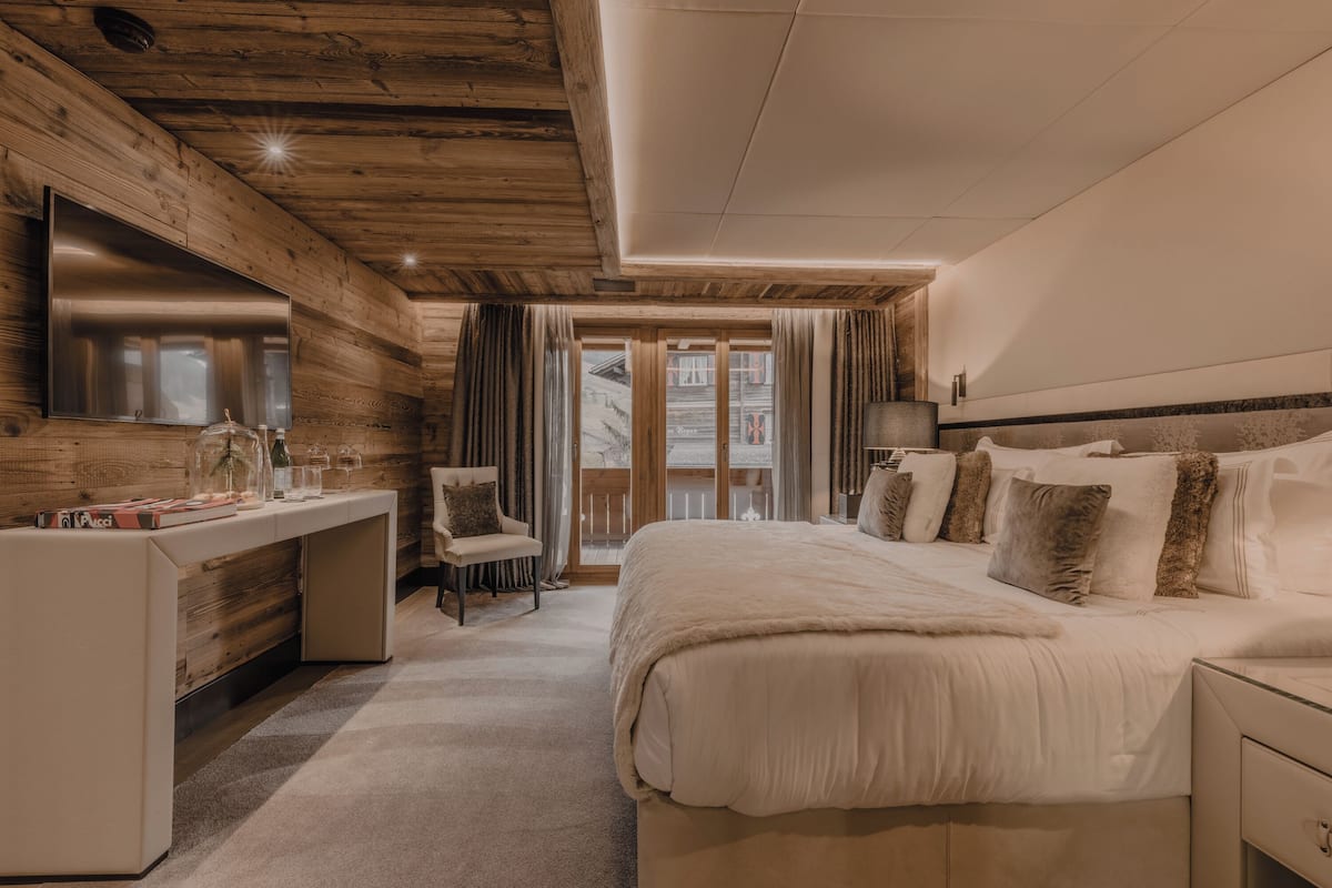 A modern hotel room with a wooden ceiling, a large bed with multiple pillows, a desk with a computer, and a large window with curtains.