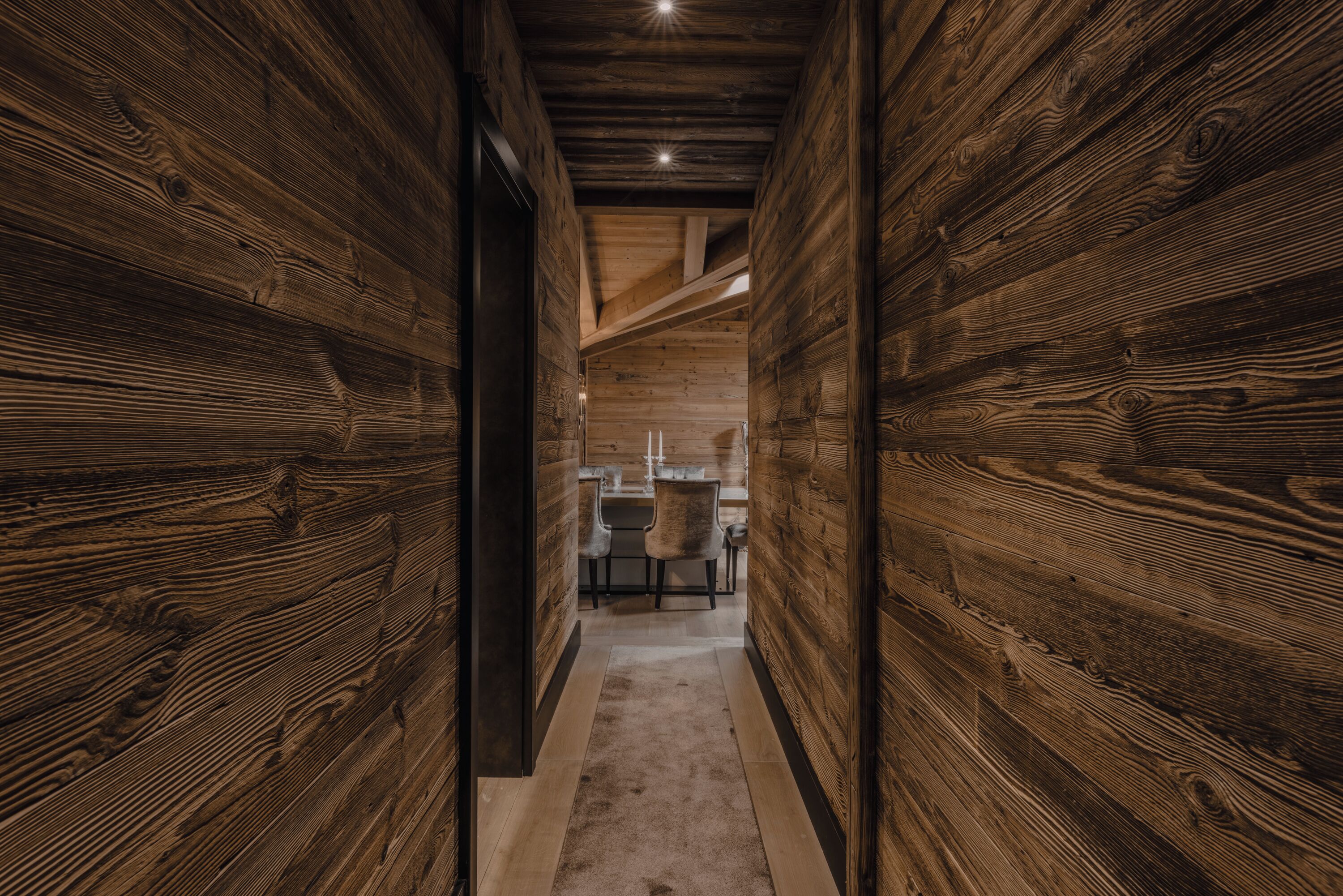 A narrow hallway with wooden walls and a carpeted floor, leading to a kitchen area with modern appliances and chairs.