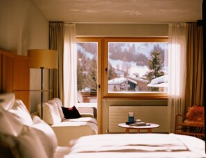 Habitación de hotel con cama, sofá, una mesita y ventana con vistas a casas cubiertas de nieve.