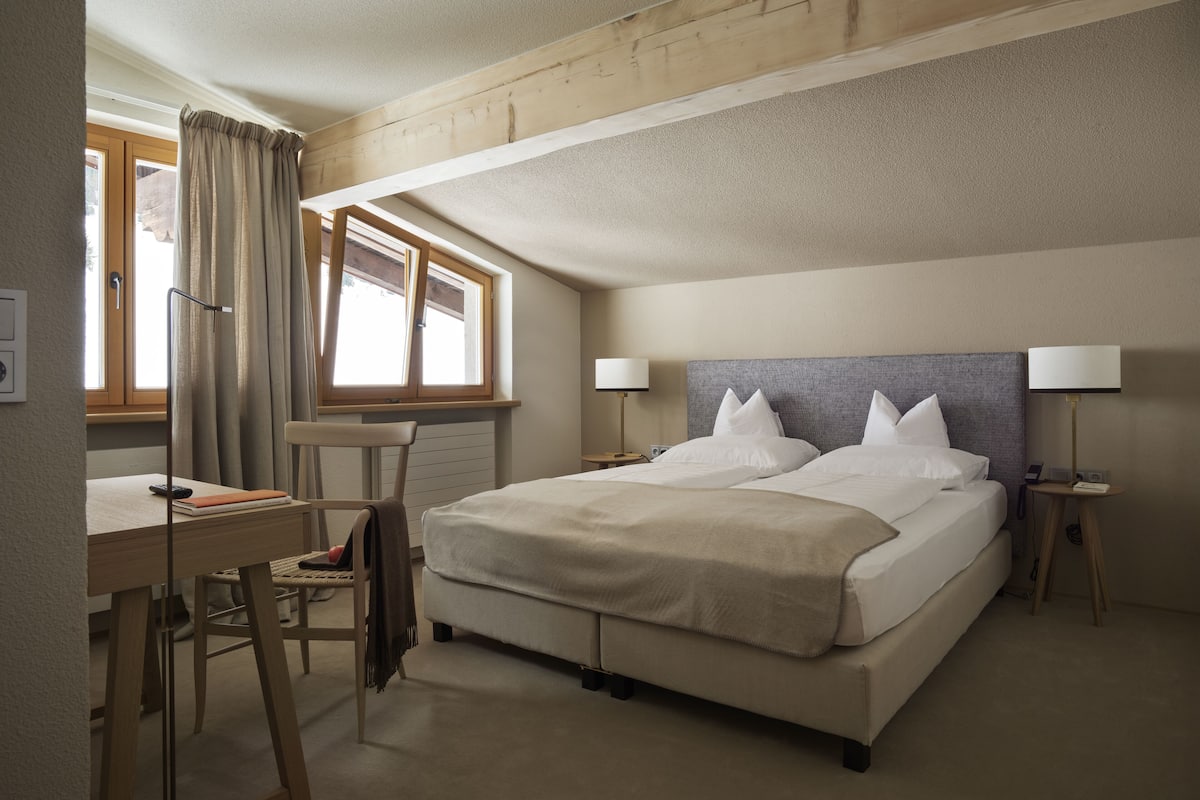 A bedroom with a bed, two bedside tables with lamps, a desk with a chair, a window with curtains, and a radiator.