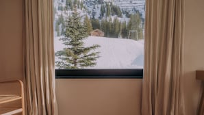 Uma janela com cortinas, uma cadeira de madeira e vista para uma montanha nevada com uma cabana.