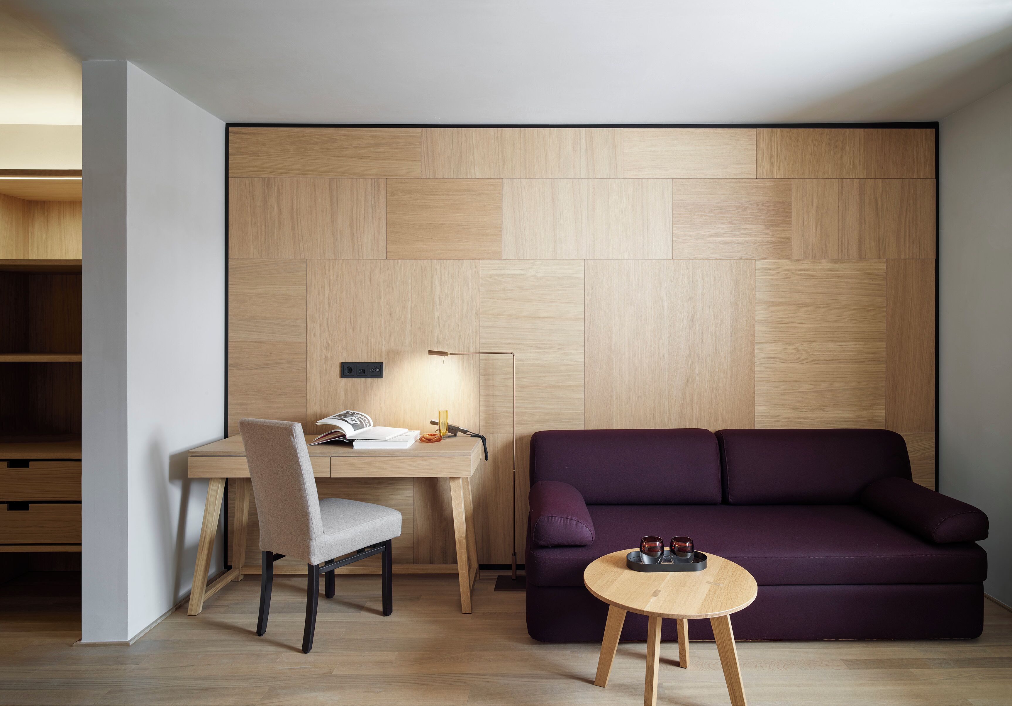 A modern room with a wooden wall panel, a desk with a chair, a purple sofa, and a small wooden table.