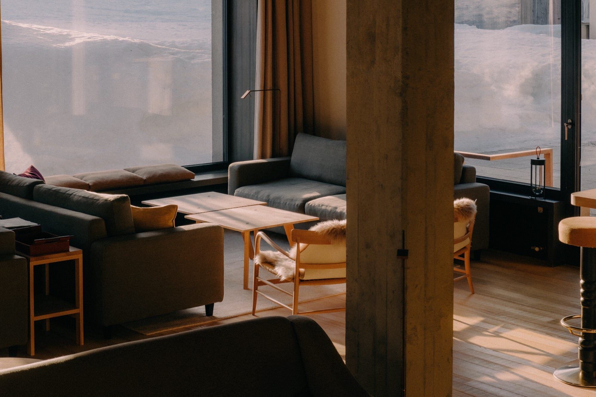 Un salon moderne avec un canapé, des fauteuils, une petite table et une vue sur un paysage enneigé, visible à travers de grandes fenêtres.