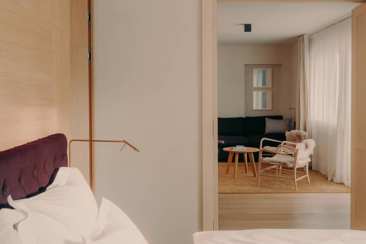 A hotel room with a bed, a wooden door, and a view into a living area with a sofa, chairs, and a teddy bear.