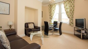 A living room with a brown sofa, a glass coffee table, a dining area with a table and chairs, a TV on a stand, and a chandelier. - Sherborne House (Basingstoke)