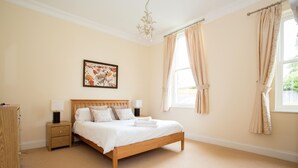 A bedroom with a wooden bed, bedside tables, a nightstand, a chandelier, and a window with curtains. - Sherborne House (Basingstoke)