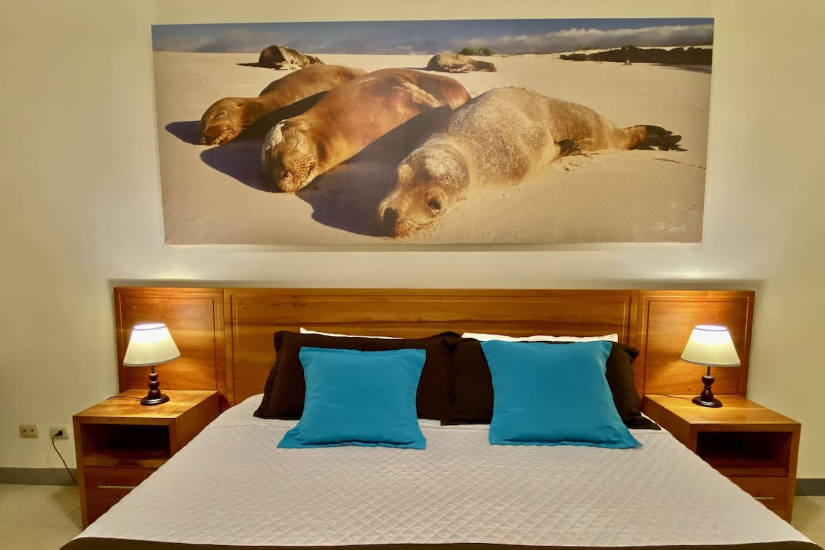 A bed with a wooden headboard, two blue pillows, and a white quilt. There are two bedside tables with lamps. Above the bed, there is a large painting of seals on a beach.