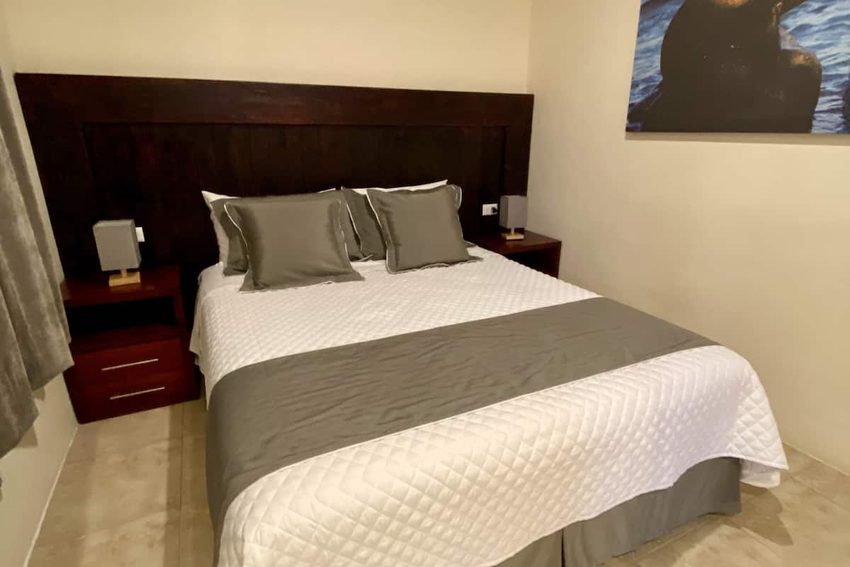 A neatly made bed with a white quilt and gray pillows, a wooden headboard, a nightstand with a lamp, and a painting on the wall.